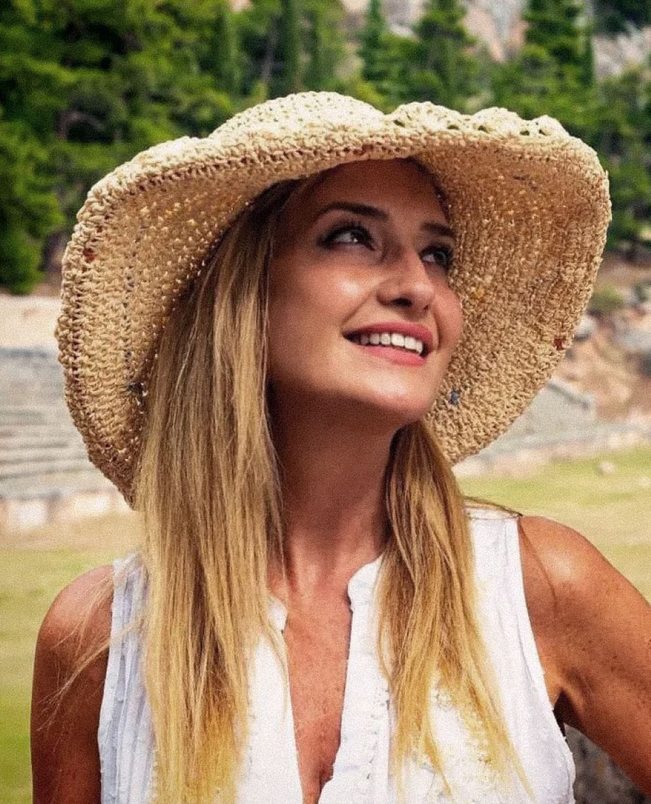 Smiling woman wearing a wide-brimmed straw hat and white striped top outdoors with green trees in the background.