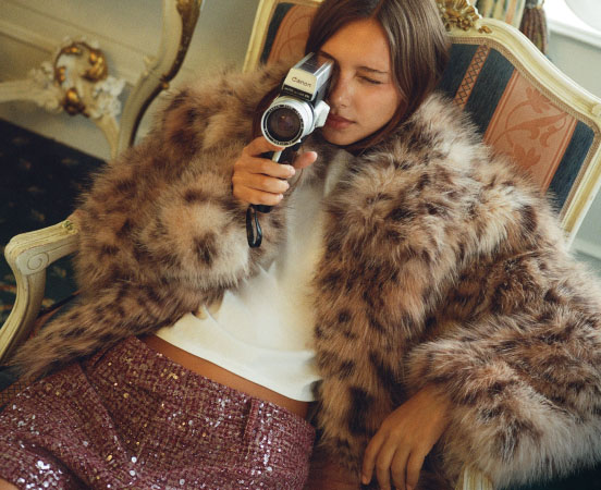 Woman in brown fur coat and sequined skirt holding a silver video camera while sitting on an ornate chair.