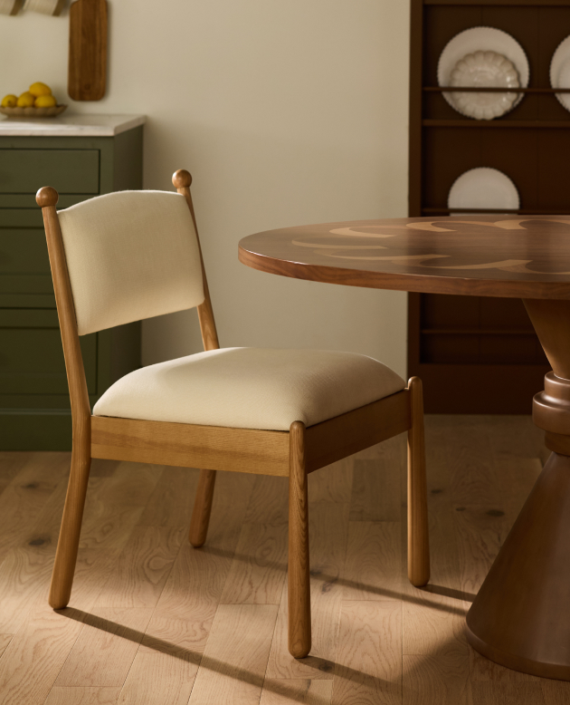 Wooden dining chair with cream upholstered seat and back beside a round wood table in a warm-toned dining room.