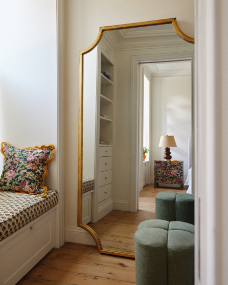 Gold-framed floor mirror reflecting a bedroom with built-in shelving, floral pillow, and green ottoman on hardwood floors.