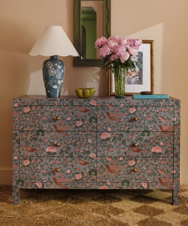 Floral-patterned dresser with pink and green design, topped with blue lamp, pink hydrangeas, and framed artwork against peach wall.
