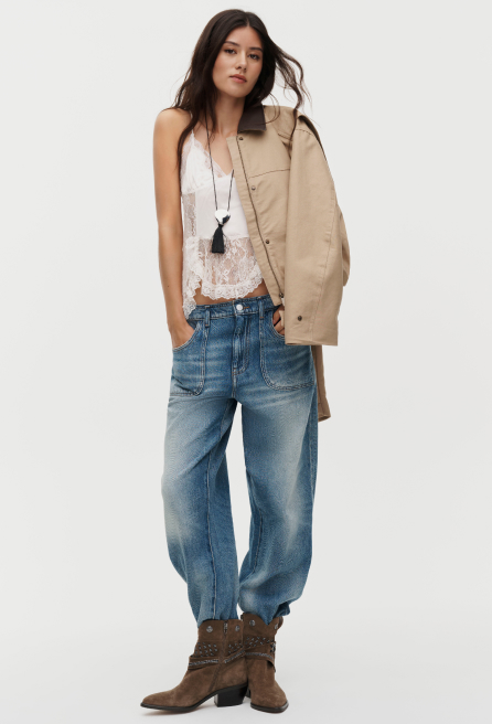Woman in white lace top, loose blue jeans, and brown boots holding a tan jacket over her shoulder on white background