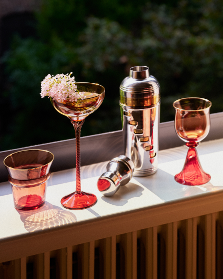 Elegant rose gold and red cocktail glasses with flower garnish, silver cocktail shaker, and jigger on white tray outdoors.