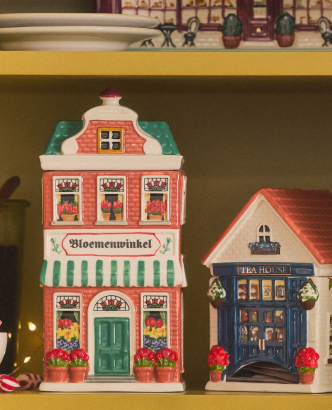 Two decorative miniature buildings on a yellow shelf: a pink Bloemenwinkel flower shop and a blue Tea House with festive details.