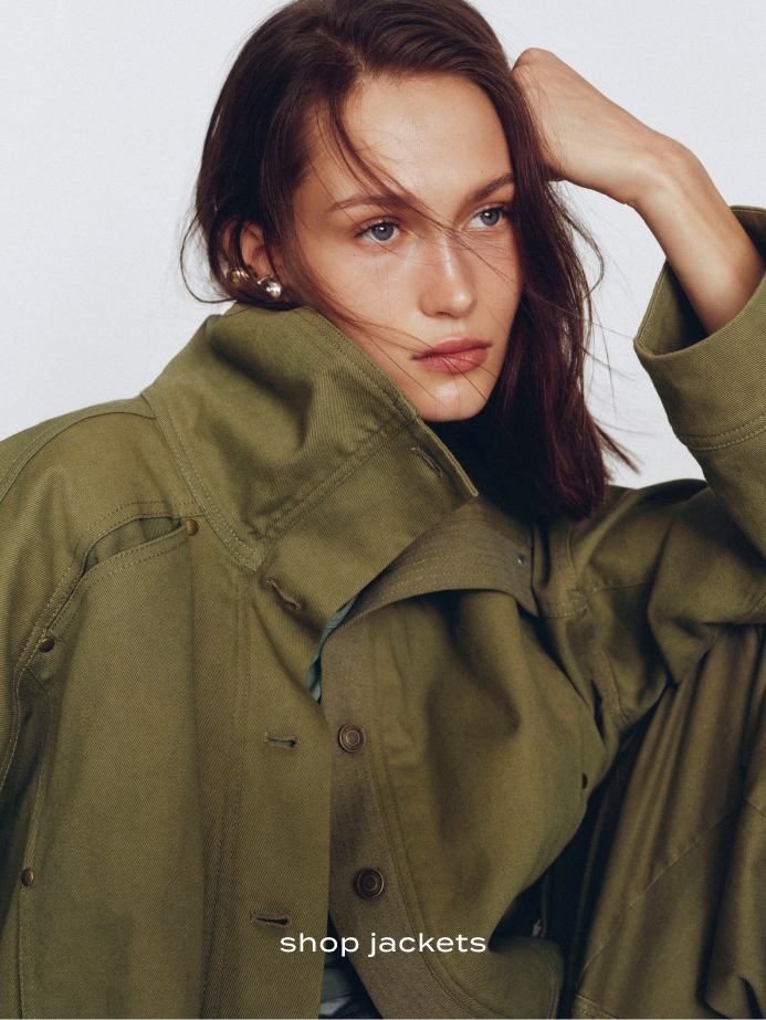 Woman with brown hair wearing olive green jacket with hand in hair against white background with text shop jackets