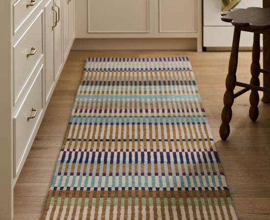 Colorful striped runner rug with horizontal bands in blue, tan, mint, and brown on hardwood floor between white cabinets and stool.