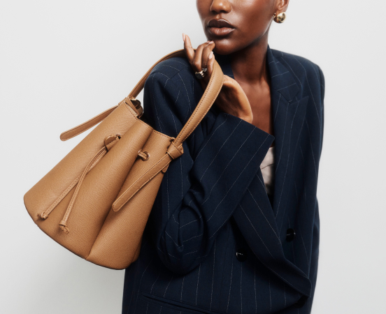 Woman in navy pinstripe blazer holding tan leather bucket bag over shoulder against light background