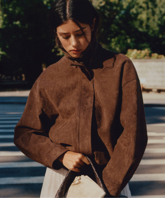 Young woman with dark hair wearing a brown jacket, looking down while holding a white handbag outdoors in sunlight.