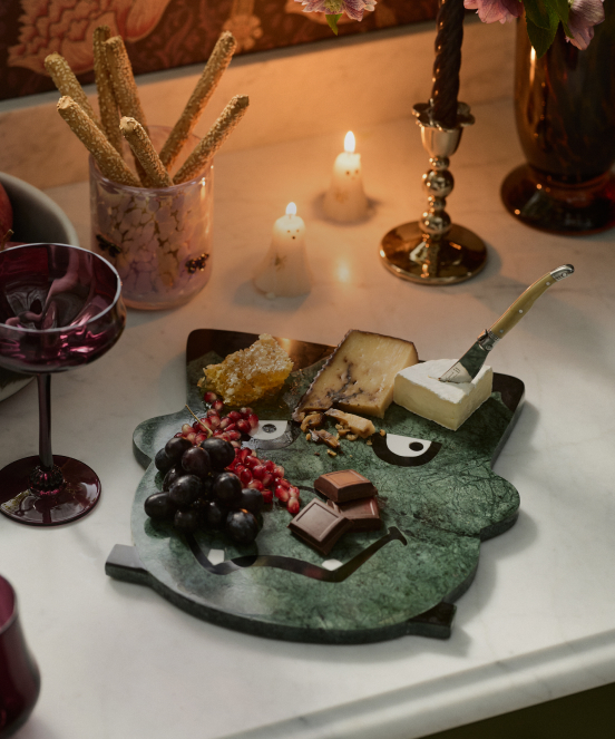 Green marble cheese board with grapes, chocolate, honeycomb, and cheese, surrounded by wine glasses, candles, and breadsticks.