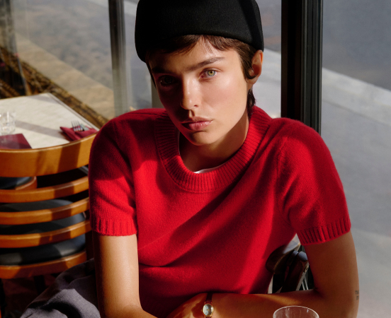 Person wearing black beanie and red short-sleeve sweater sitting at table with glass, natural lighting from window