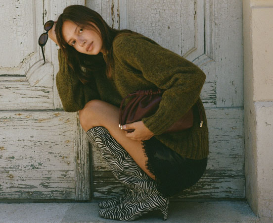 Woman in olive green sweater and burgundy shorts crouching against weathered white wooden door wearing zebra-print boots