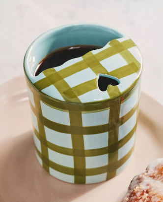 Coffee cup with green and white checkered pattern lid on a peach-colored surface with crumbs nearby.