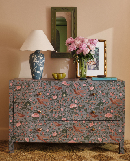 Floral-patterned dresser with blue lamp, pink flowers in vase, framed art, and green mirror against peach wall.