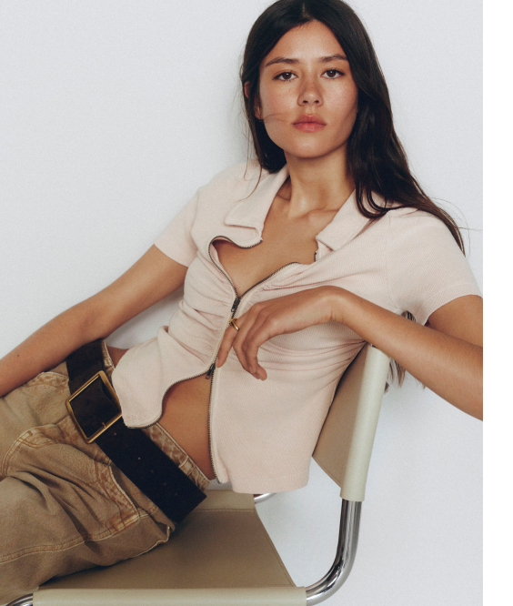 Woman with long dark hair wearing beige short-sleeve shirt and tan pants, sitting on white chair against white background.