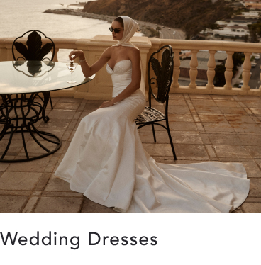 Bride in elegant white wedding dress seated at outdoor terrace table overlooking coastal view with text "Wedding Dresses"
