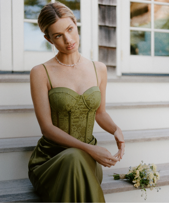 Woman in olive green corset top and skirt sitting on white steps with a small bouquet, wearing a delicate necklace.
