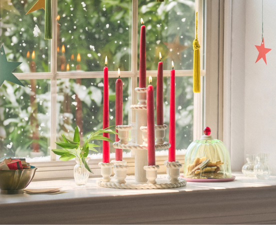 Red and white candles in holders on windowsill with snowy Christmas tree visible through window and holiday decorations