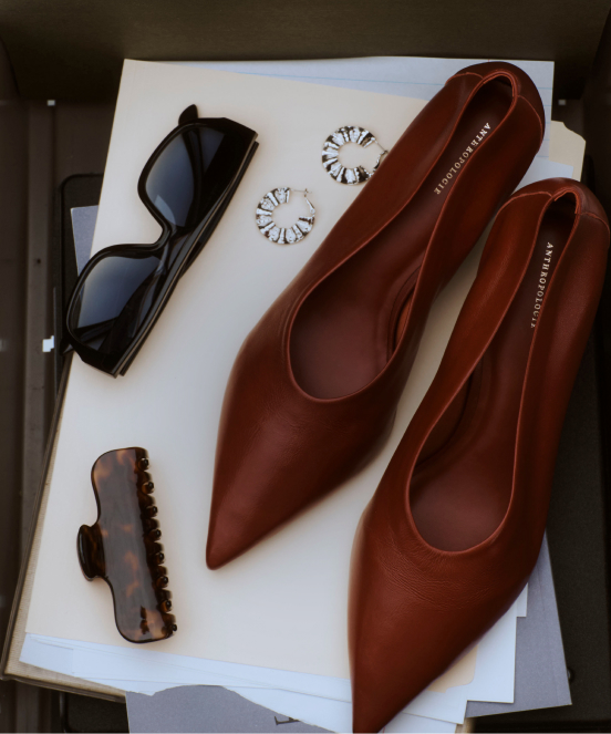 Brown leather pointed-toe flats on white papers with sunglasses, daisy earrings, and a brown hair clip arranged nearby.