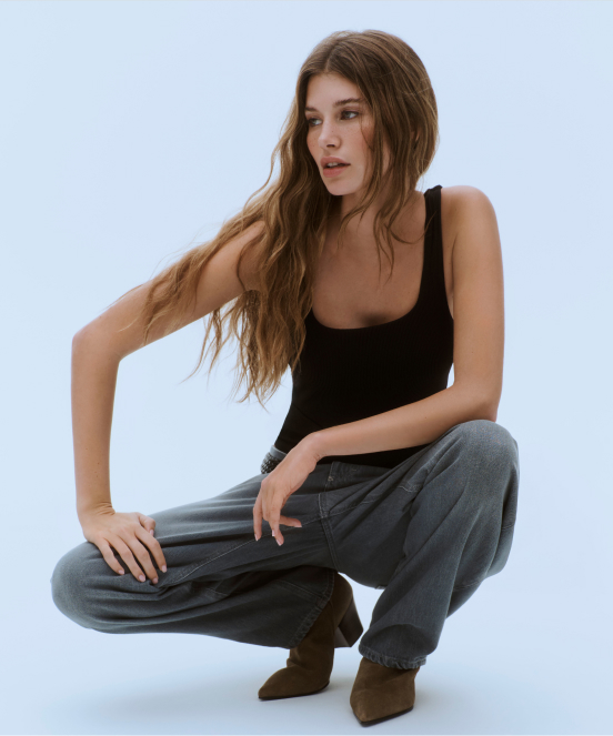 Woman with long brown hair crouching in black tank top, gray sweatpants, and brown suede shoes against light blue background.