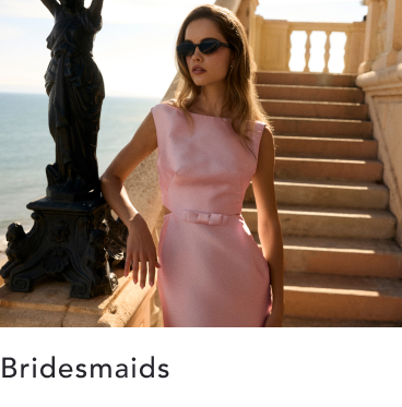 Woman in pink sleeveless dress and sunglasses standing by ornate black railing with ocean and stairs in background