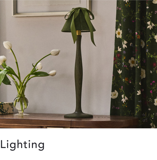 Green decorative table lamp with leaf-shaped shade on wooden surface, white tulips in vase nearby, floral curtain backdrop