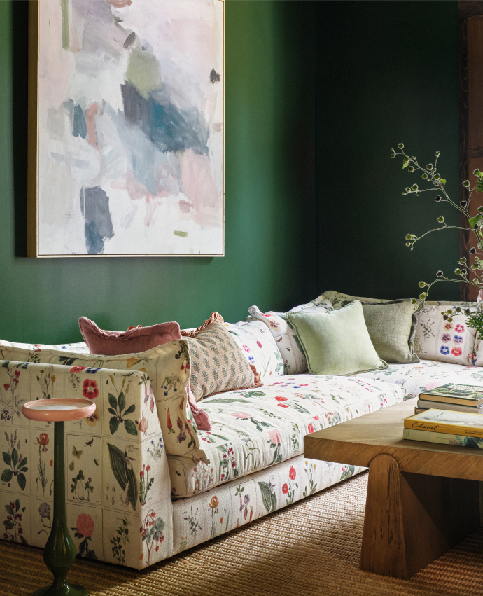 Floral-patterned sofa with colorful pillows against a dark green wall, abstract painting above, wooden coffee table nearby.