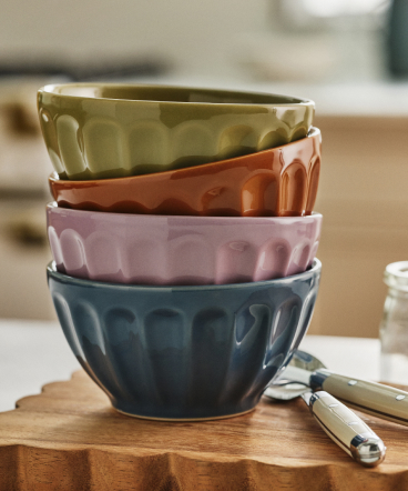 Stack of four ceramic mixing bowls in green, orange, pink, and blue with fluted edges on a wooden cutting board