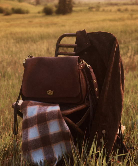 Brown leather bags and plaid blanket arranged on a folding chair in a golden grass field with blurred landscape background.