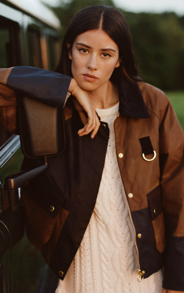 Woman with long dark hair wearing brown and black jacket with gold hardware over cream sweater, leaning against vehicle outdoors.