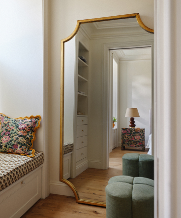 Gold-framed mirror reflecting a bedroom with built-in white shelving, floral pillow, and green ottoman on hardwood floor.
