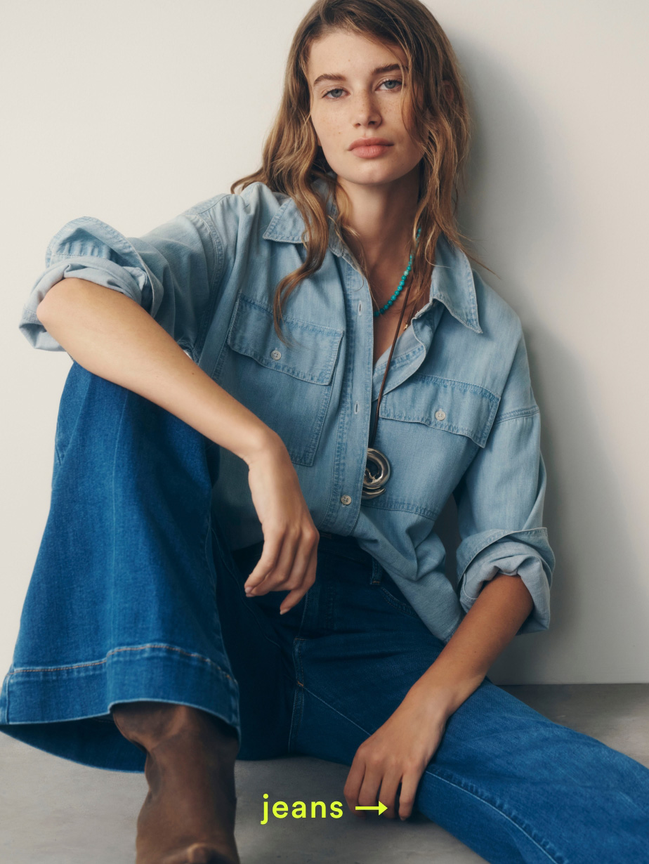Woman in light blue denim shirt and blue jeans sitting against white wall with yellow text reading "jeans" at bottom.