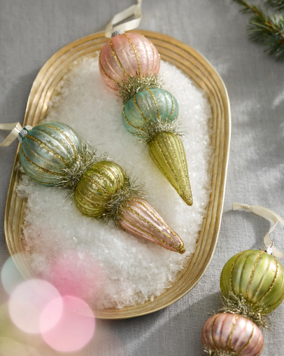 Pastel pink, green, and gold glittered ornaments arranged on a gold tray filled with white snow on gray linen background.