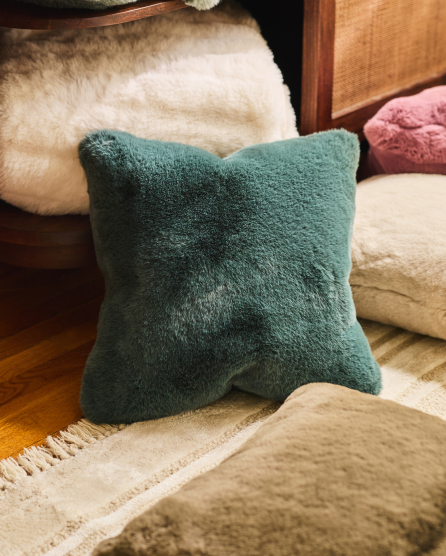 Fluffy teal green pillow on cream rug with wooden furniture and white sheepskin in background on hardwood floor
