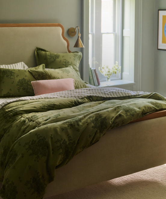 Cozy bedroom with olive green velvet bedding, pink accent pillow, wooden headboard, wall sconce, and gray-green walls.
