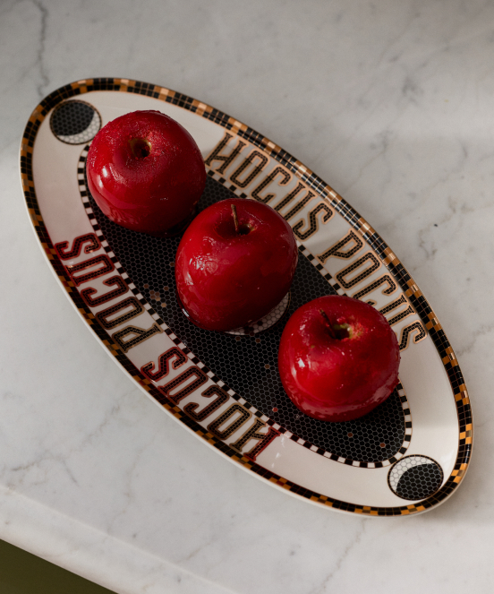 Three glossy red apples on an oval decorative tray with text reading "HOCUS POCUS" and "FOCUS SCHMOCUS" on a marble surface.