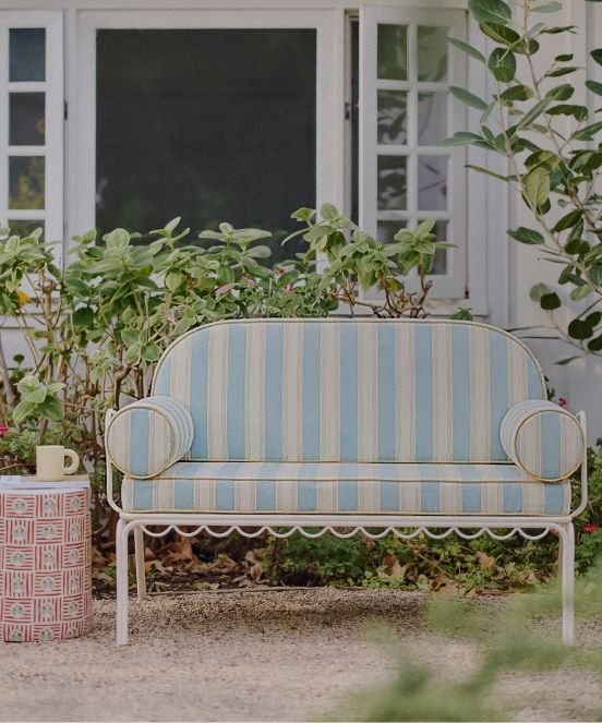 Blue and beige striped cushioned outdoor bench with white metal frame on patio with plants and white house entrance.