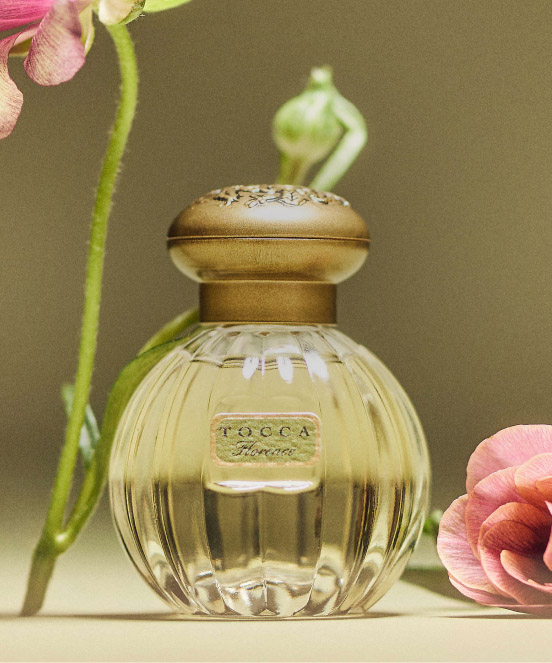 Tocca Florence perfume bottle with gold cap and ribbed glass design, surrounded by pink flowers on neutral background.