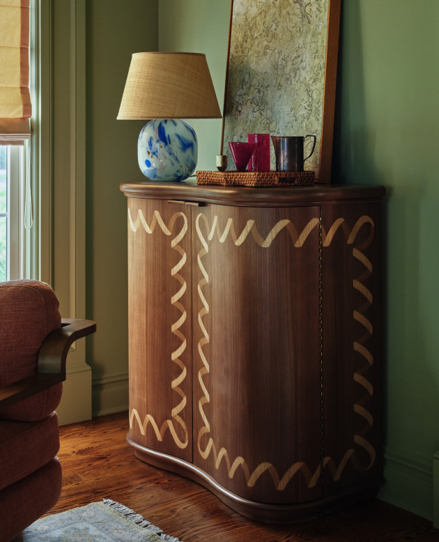 Wooden cabinet with decorative zigzag pattern, topped with blue and white lamp, wicker tray, and mugs against green wall