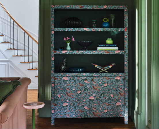 Built-in shelving unit with floral wallpaper backing and green interior, displaying books and decorative objects beside staircase.