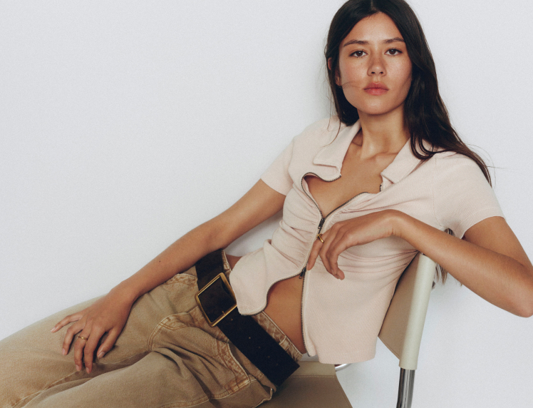 Woman with long dark hair wearing a beige tied shirt and tan pants with black belt, sitting on white chair against white background.
