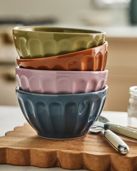Stack of four ceramic bowls in green, orange, pink, and blue with fluted edges on a wooden cutting board with utensils nearby.