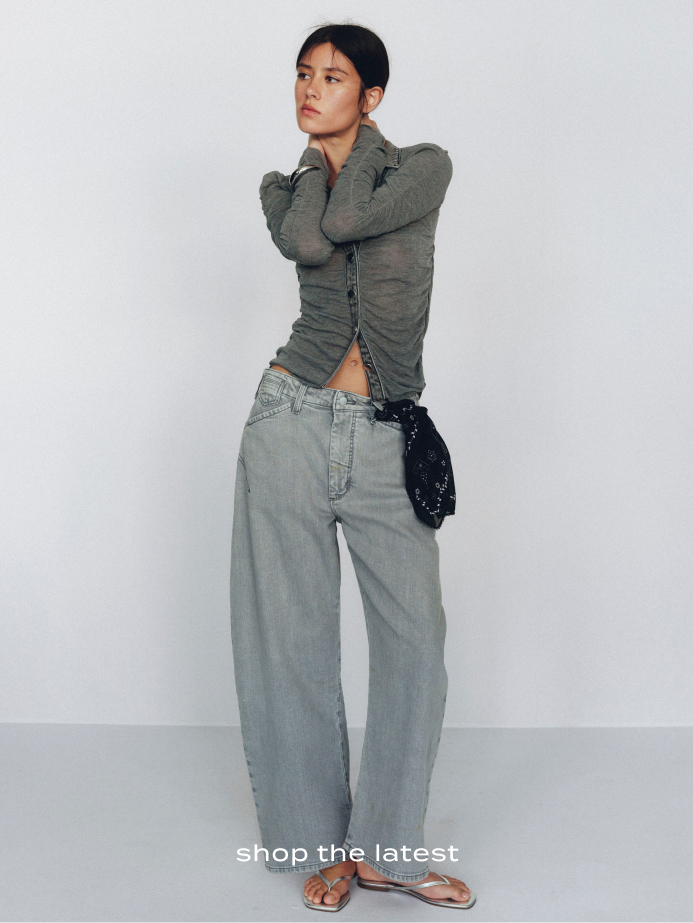 Woman in gray ruched long-sleeve top and wide-leg jeans holding a black bag against white background with text shop the latest