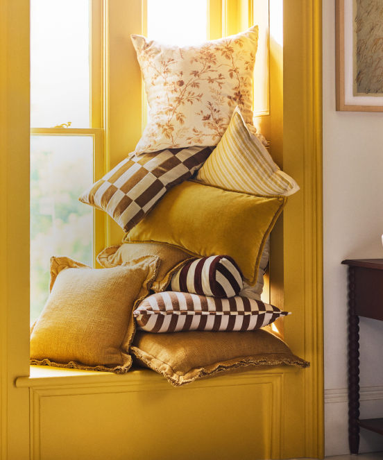 Stack of decorative throw pillows in yellow, gold, and brown tones with various patterns arranged on a yellow window seat.