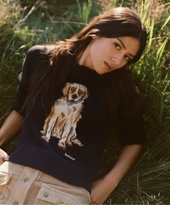 Woman in black Barbour sweater with dog graphic lying in tall grass, looking at camera with natural sunlight.