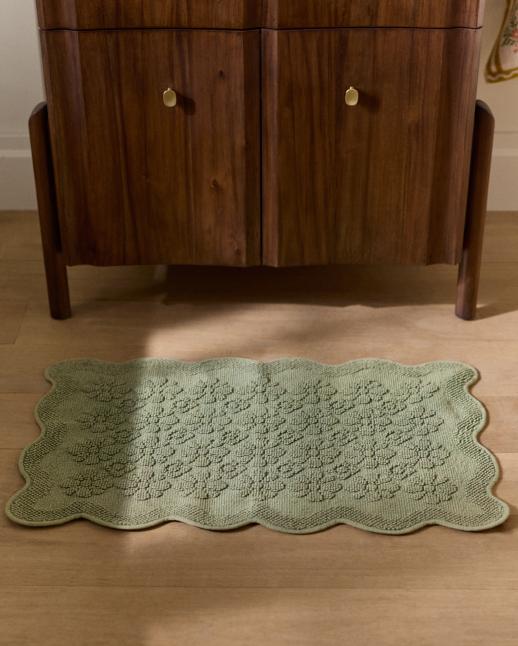 Sage green textured bath mat with scalloped edges on wooden floor in front of walnut cabinet with brass knobs