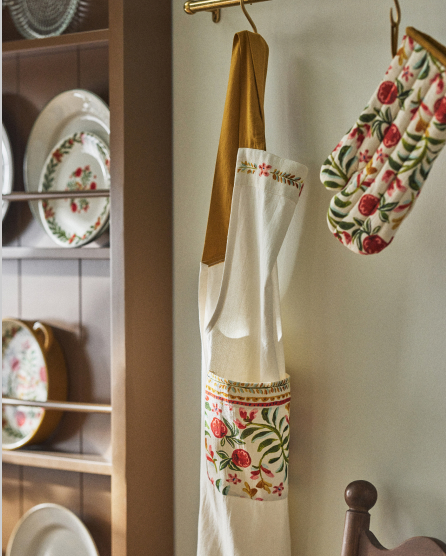 White apron with floral trim and pocket hanging on hook, with matching dishes displayed on shelves in background.