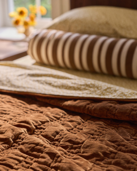 Stack of folded textured blankets and throws in earth tones including rust, beige, and brown stripes with yellow flowers in background