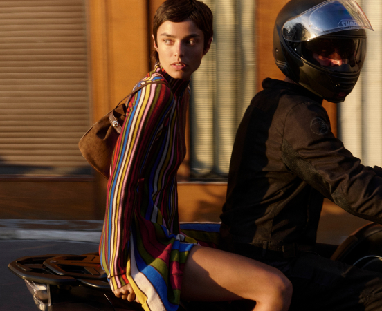 Woman in colorful striped shirt sitting on motorcycle behind person wearing black helmet and jacket in warm-lit room.