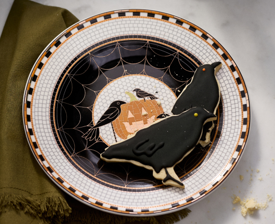 Halloween plate with black cat cookie, pumpkin, ravens, spider web design, and orange and black checkered border on white background.