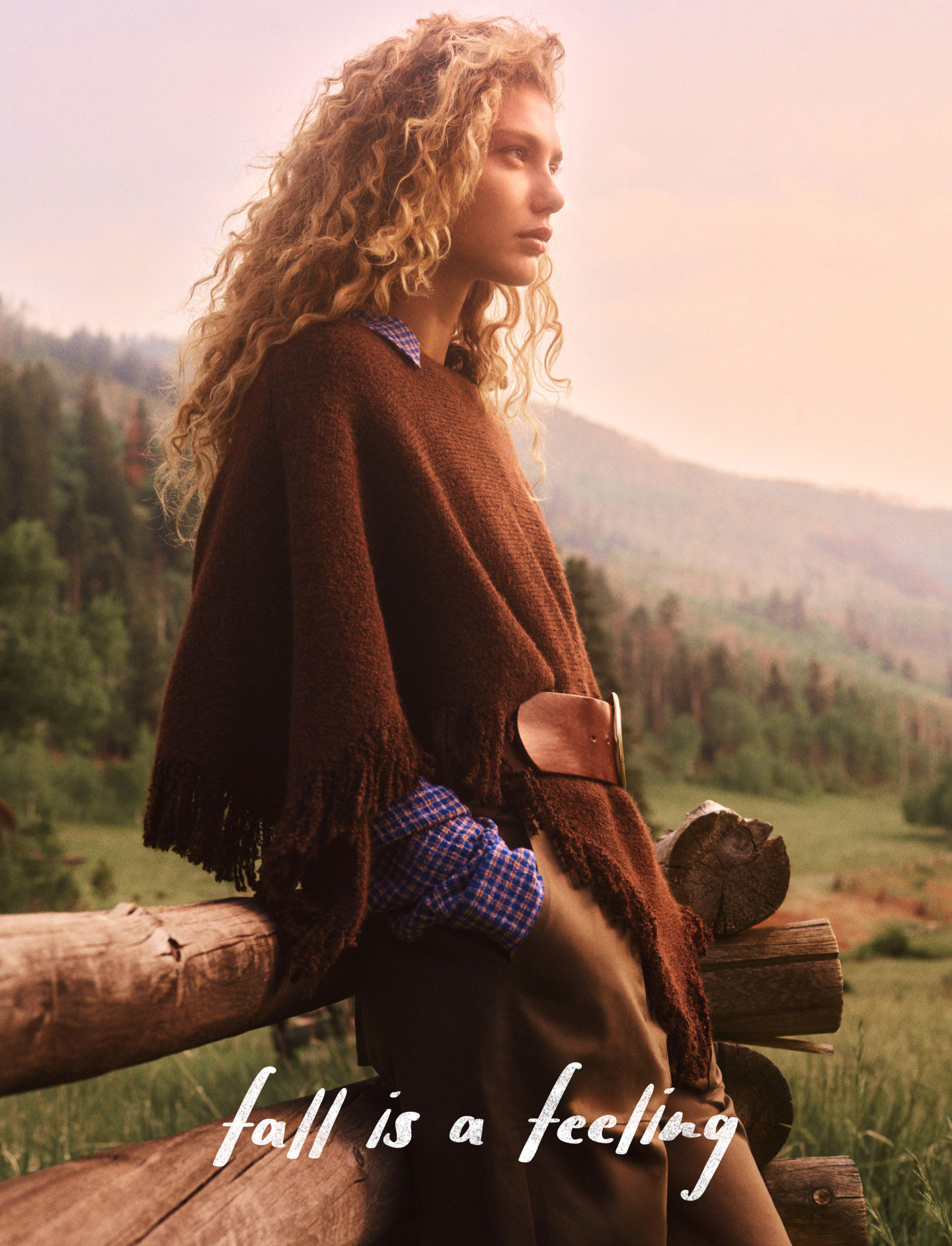 Woman with curly blonde hair wearing brown poncho sits on wooden fence overlooking mountain landscape. Text reads fall is a feeling.